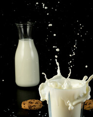The milk is splashing out of the glass, creating a mess on the table. A cookie is also present on the table, adding to the scene. Black background