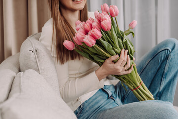 Young woman holding a bouquet of pink tulips, sitting on a cozy couch. Peaceful and thoughtful expression. Soft natural light, modern home interior, spring vibes. 8 March.