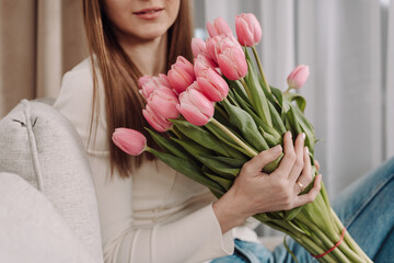 Young woman holding a bouquet of pink tulips, sitting on a cozy couch. Peaceful and thoughtful expression. Soft natural light, modern home interior, spring vibes. 8 March.