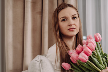 Young woman holding a bouquet of pink tulips, sitting on a cozy couch. Peaceful and thoughtful expression. Soft natural light, modern home interior, spring vibes. 8 March.