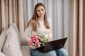 Young woman with a laptop sitting on a cozy couch, looking away thoughtfully. Pink tulips beside her. Modern home interior, relaxed atmosphere. Remote work, freelance lifestyle.