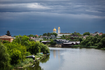 Fototapeta premium A picturesque town with a river in the foreground, lush greenery, and golden-domed churches in the background. The dramatic sky adds contrast, creating a serene yet moody atmosphere.