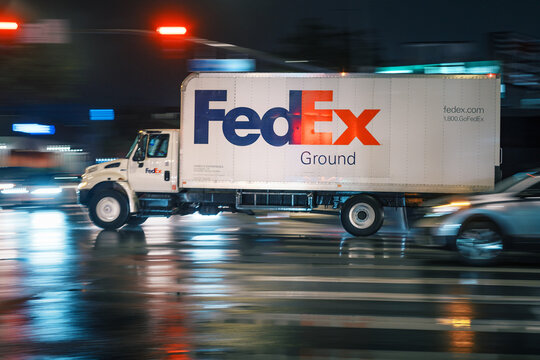 LOS ANGELES, CA - March 07, 2025: FedEx delivery truck rushing through city traffic, crossing illuminated street intersection on a rainy night in West Hollywood, California.