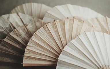 Close-up view of a collection of delicate, folded paper fans in various shades of beige, cream, and off-white. The fans are arranged overlapping each other, creating a textured and visually appealing