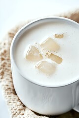 Fresh milk or cream in white ceramic cup with melting ice cubes creating natural patterns on surface, placed on knitted textile background.