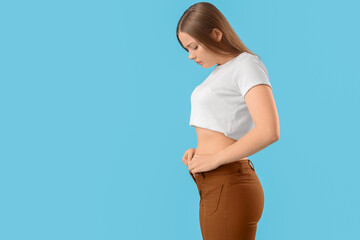 Young woman trying to button tight pants on blue background. Weight gain concept