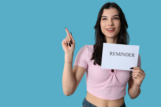 Pretty young woman holding paper sheet with text REMINDER and pointing up on blue background