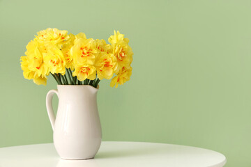 Daffodil flowers in jug on coffee table against green background