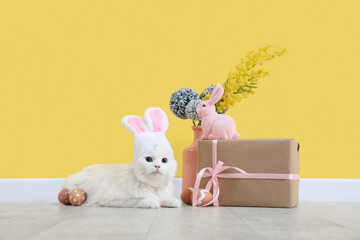 Cute white cat in bunny ears with Easter eggs, gift box, toy rabbit and beautiful flowers near...