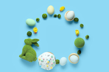 Frame made of green flowers, decor, Easter eggs and cake on blue background