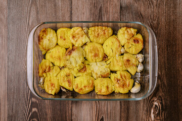 Baked golden potatoes with garlic cloves lie in a glass dish on the table. Top view