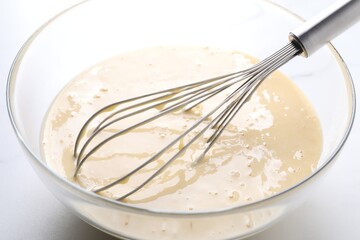 Whisk and bowl of dough on white table, closeup