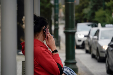Young woman in red hoodie, engaged in a phone call, leaning against a pole, with urban street and traffic in the background
