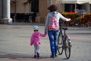 Fototapeta premium Mother and daughter walking hand-in-hand with a bicycle, enjoying a sunny day in an inviting urban environment