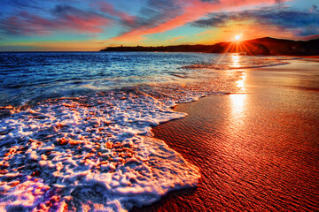 A serene beach scene at sunrise, featuring gentle waves lapping the shore with golden sunlight reflecting on the sand. The colorful sky and calm ocean create a peaceful atmosphere, ideal for travel, r