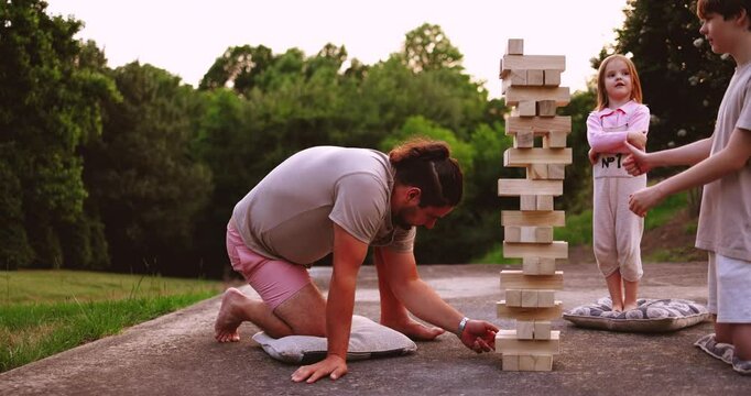 Family Playing Wooden Block Tower Game on summer day during sunset. Fun Family Outdoor Game. The Moment of Tumbling Block Tower Collapsing