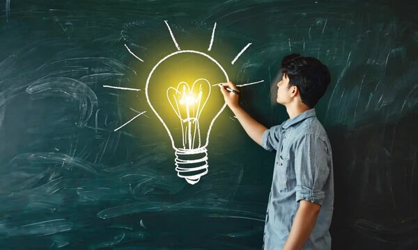 A young man sketching a glowing lightbulb on a chalkboard in a classroom setting.