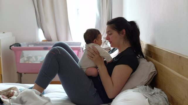 Mother lying on bed while holding her baby, sharing a tender kiss with natural daylight through window in the background. Cozy home environment with soft light curtains and pink baby bassinet