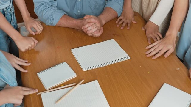 Paper scatter on table while caucasian teacher and diverse children putting hand together to celebrate successful project. Represent love, corporate, team working, togetherness and unity. Edification