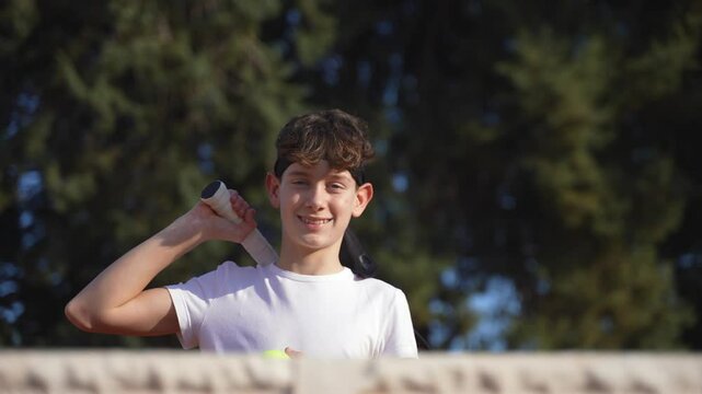 A happy teenage male tennis player standing behind the net, picks up the racket and the ball and smiling at the camera.