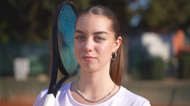 Closeup portrait of a happy teenage girl tennis player standing behind the net, picks up the racket and the ball and smiling at the camera.