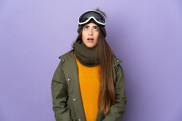 Skier caucasian girl with snowboarding glasses isolated on purple background looking up and with surprised expression