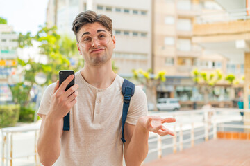 Young handsome man using mobile phone at outdoors making doubts gesture while lifting the shoulders