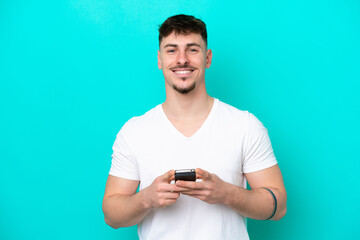 Young caucasian handsome man isolated on blue background looking at the camera and smiling while using the mobile