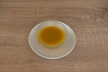 A small bowl of yellow liquid sits on a white plate on a wooden table