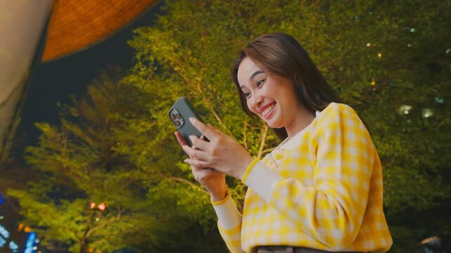 Smiling woman in yellow sweater joyfully using smartphone outdoors at night, surrounded by trees, embracing happiness and casual moments under stars. businesswoman casual