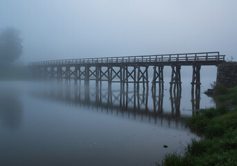 Fototapeta premium A rustic wooden bridge over a serene, fog-covered river.