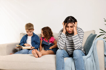 Overwhelmed mother and her naughty children with book on sofa at home