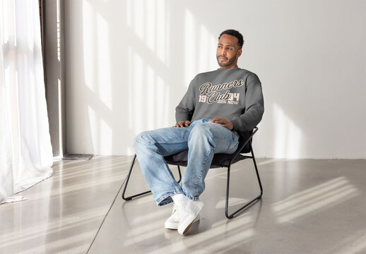 A black man sits in a minimalist room, wearing a stylish sweater and jeans. PSD mockup template