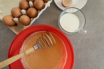 A red mixing bowl contains flour and a cracked egg, with a whisk resting inside.