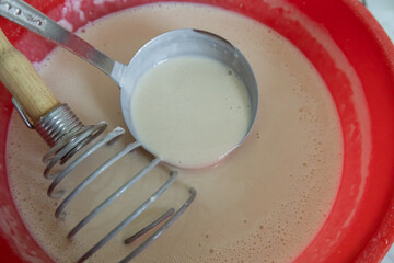 A close-up view of a smooth pancake batter mixture in a red bowl. A metal ladle is lifting some batter, while a spiral whisk with a wooden handle rests inside the bowl. 