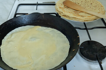 A thin crepe is cooking in a black frying pan on a gas stove, with a stack of prepared crepes on a plate in the background. 