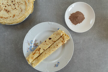 Two homemade crepes are rolled and placed on a vintage floral plate, with a stack of additional crepes in the background. 