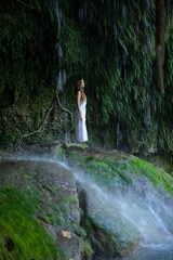 Naklejka premium A serene woman in a white dress stands under the cascading Antalya Famous Duden Waterfall, surrounded by moss-covered rocks, lush greenery, and flowing water in a peaceful natural setting. 