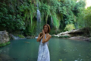 Fototapeta premium A joyful woman in a white dress stands near a turquoise river with a waterfall in the background, surrounded by lush greenery, expressing happiness and serenity. 