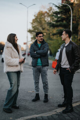 Three young adults are seen conversing casually outdoors, dressed comfortably for cool weather....
