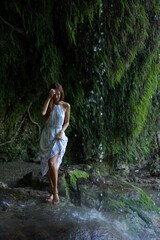 Fototapeta premium A woman in a white dress stands near a cascading waterfall, touching her hair as she enjoys the refreshing mist, surrounded by lush greenery and moss-covered rocks. 