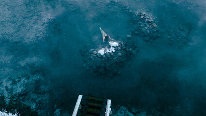 Aerial view of a lone woman relaxing in a stunning thermal spring surrounded by snowy landscapes in...