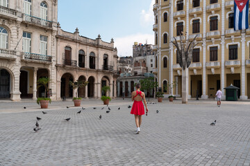 Red dressed woman strolling plaza vieja, surrounded by pigeons, colonial architecture framing...