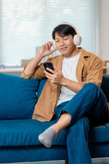 Fototapeta premium Young asian male listening to music via wireless headphones, reclining on couch with smartphone nearby