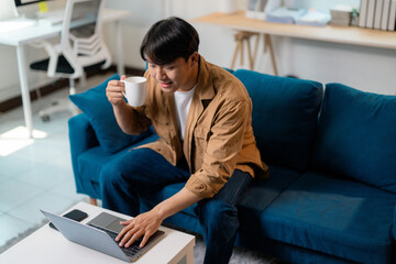 Young Asian professional sipping coffee, typing on laptop while relaxing on blue living room sofa