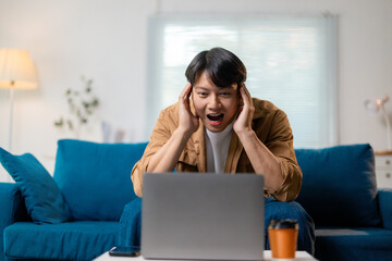 Stressed asian freelancer receiving negative email, working remotely on laptop while sitting on living room sofa