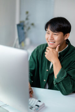 Happy young asian man working from his home office using a desktop computer