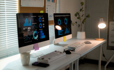 Data analysis workspace with dual monitors displaying charts, professional desk lamp highlighting modern office environment