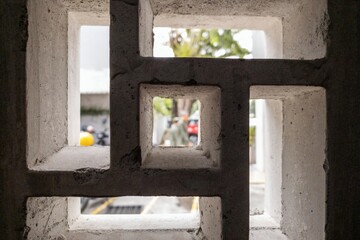 Close-up of a concrete ventilation block with geometric square patterns. The background shows a blurred street scene with a person, trees, and vehicles, framed through the openings.