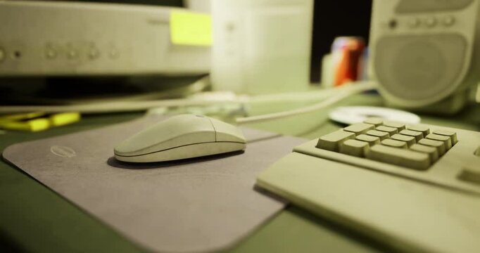 A computer mouse rests on a mouse pad beside a keyboard in a dimly lit workspace. The desk features various items including speakers and a clutter of office supplies in the background.